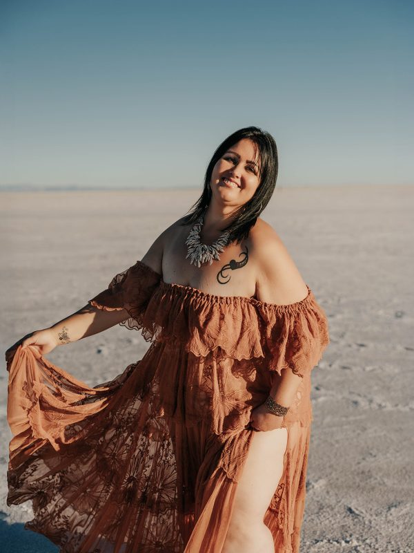 Confident plus-size woman in a flowing terracotta off-shoulder boho dress twirling and laughing on the Bonneville Salt Flats in Utah during a Kali energy women's retreat, dress by WeAreReclamation