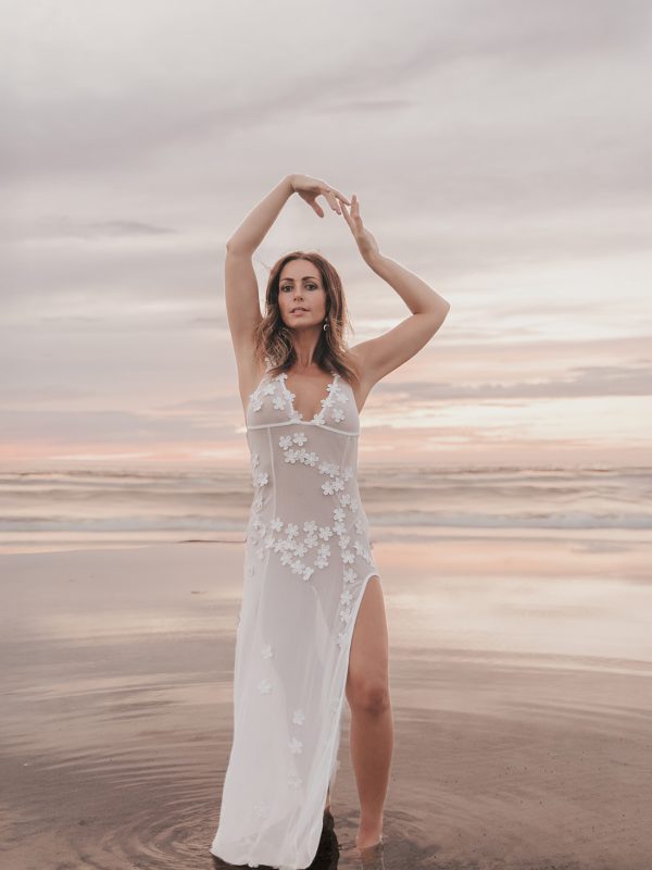 Woman in a sheer white floral dress standing in shallow water at sunset with arms raised during the Kali Experience day retreat at the Salt Lake City salt flats, embodying ego dissolution and divine feminine awakening