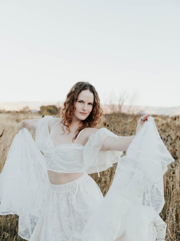 Woman in flowing white lace two-piece outfit standing in a golden Utah field during a women's retreat, arms outstretched with fabric billowing at sunset
