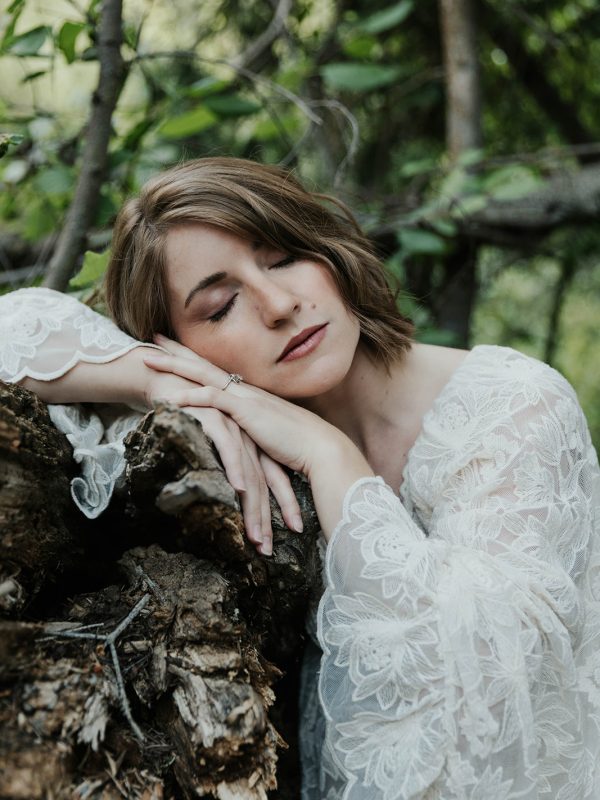 Close-up of a woman in a white lace robe resting her cheek on her hands against a mossy log in a Utah forest with eyes closed in peaceful surrender during a women's wellness retreat